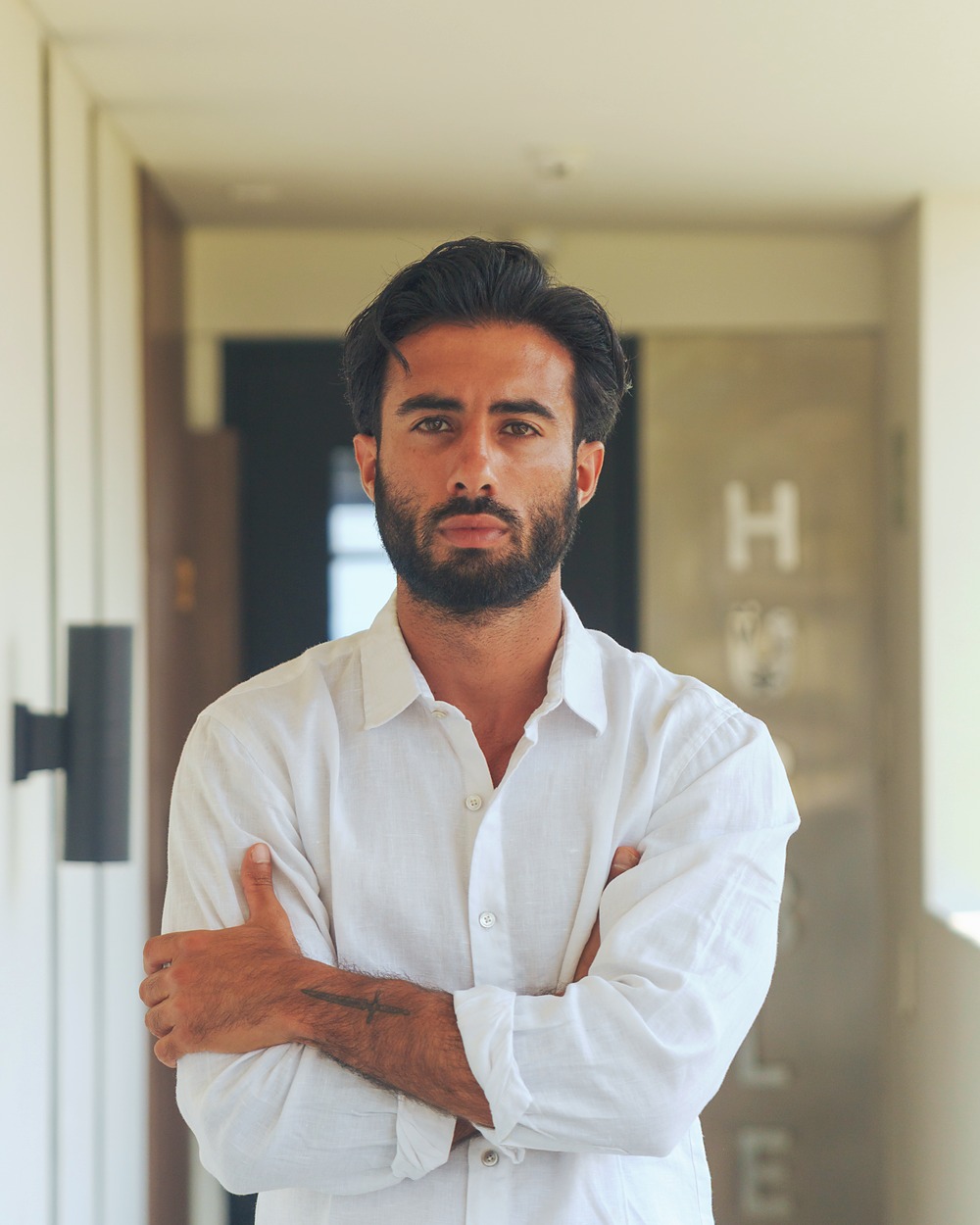 Suroesh Jazayeri standing in a warm ochre corridor, arms crossed.